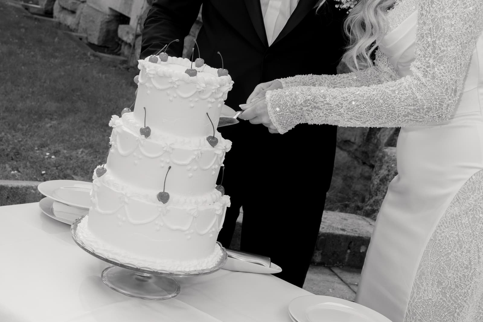 Brautpaar schneidet gemeinsam mehrstöckige weiße Hochzeitstorte mit Herzdekorationen an, Braut im spitzenverziertem Kleid, Bräutigam im schwarzen Anzug